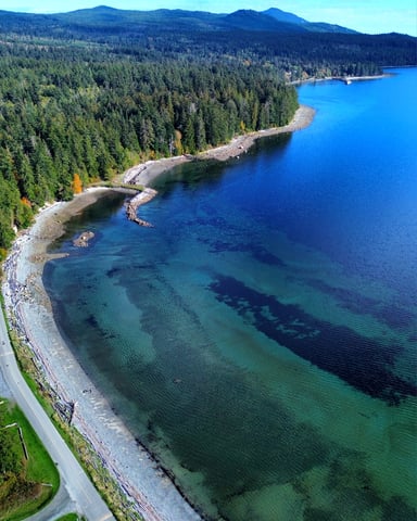 Aerial view of turquoise lake with rocky shoreline, dense coniferous forest, mountains, and coastal road