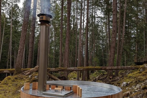 Wooden hot tub with metal rim and chimney in forest clearing surrounded by tall pine trees