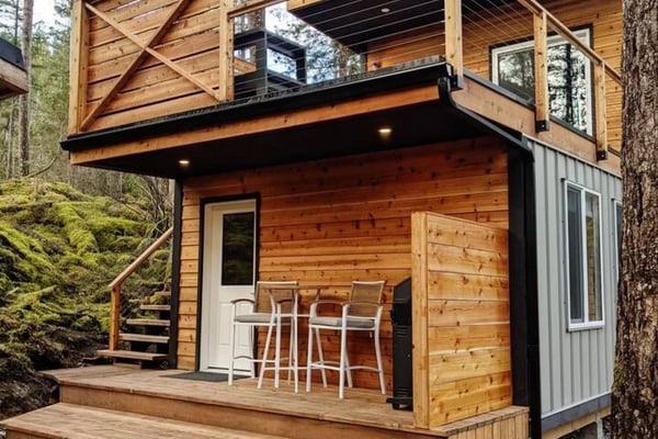 Modern wooden cabin with upper deck and metal railings nestled among tall pine trees in forest setting.