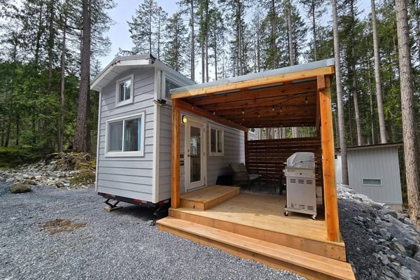 Tiny house with wooden deck and pergola, gas grill, surrounded by tall pine trees in forest setting
