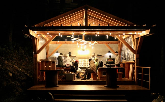 Illuminated wooden pavilion at night with guests gathered around a kitchen counter and cooking area