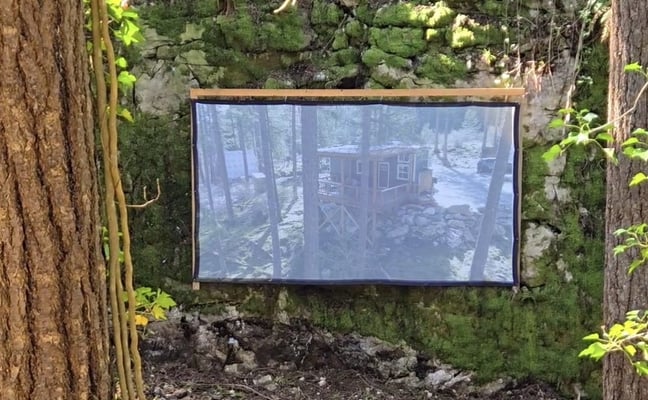 Outdoor screen mounted on moss-covered stone wall between tall trees, displaying forest cabin scene