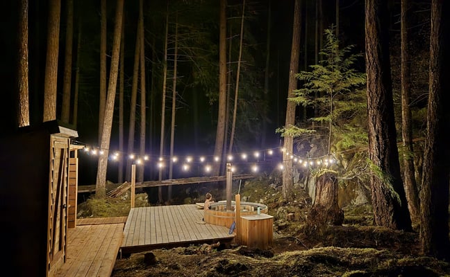 Nighttime forest deck with string lights, wooden hot tubs, and tall pine trees surrounding illuminated outdoor space