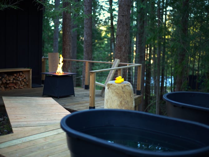 Wooden deck in forest with fire pit, soaking tubs, and tall trees in background at dusk