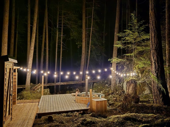Nighttime forest deck with string lights, wooden hot tubs, and tall evergreen trees